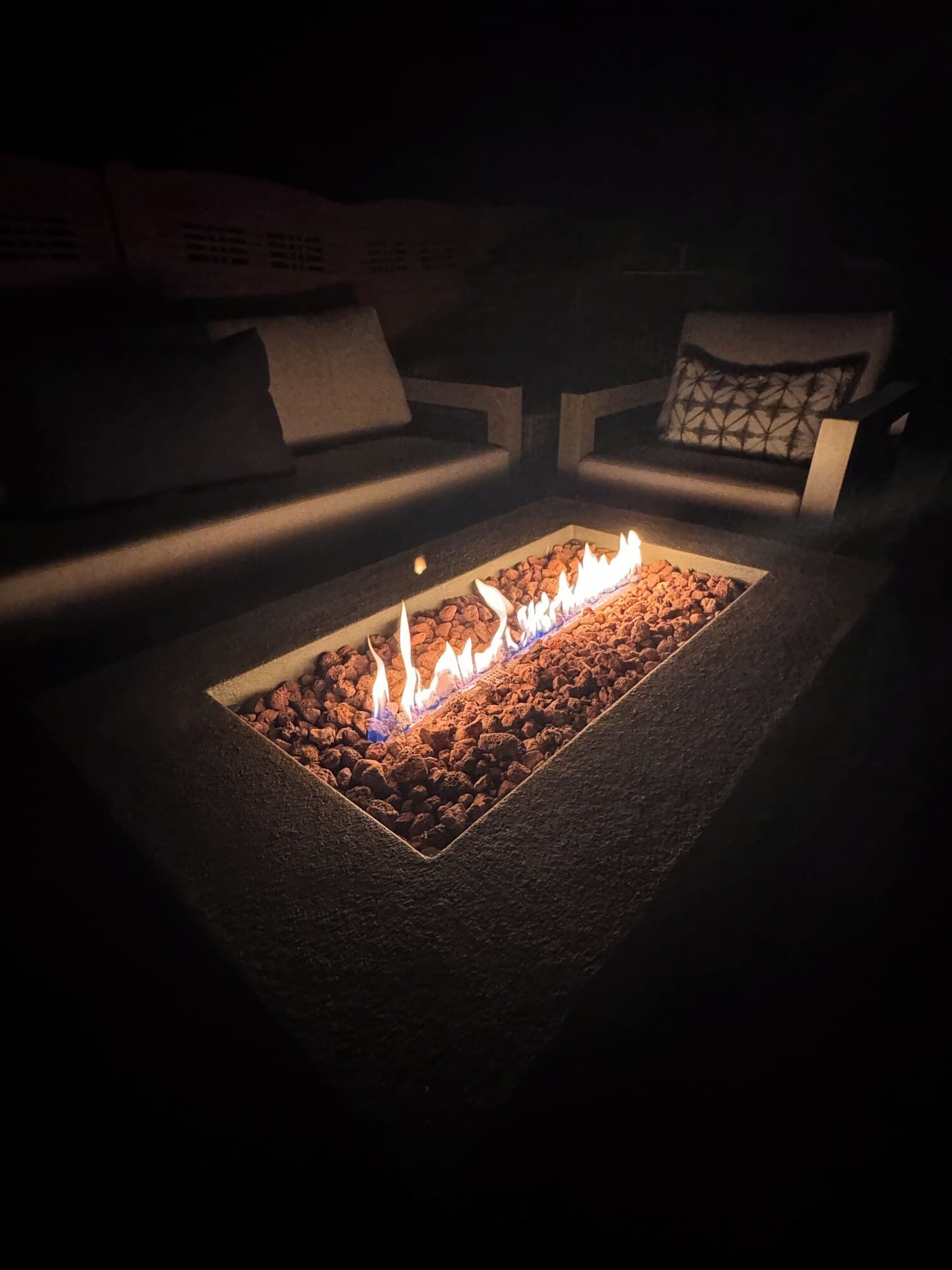 A rectangular outdoor fire pit with a fire burning in the center, surrounded by a stone border.