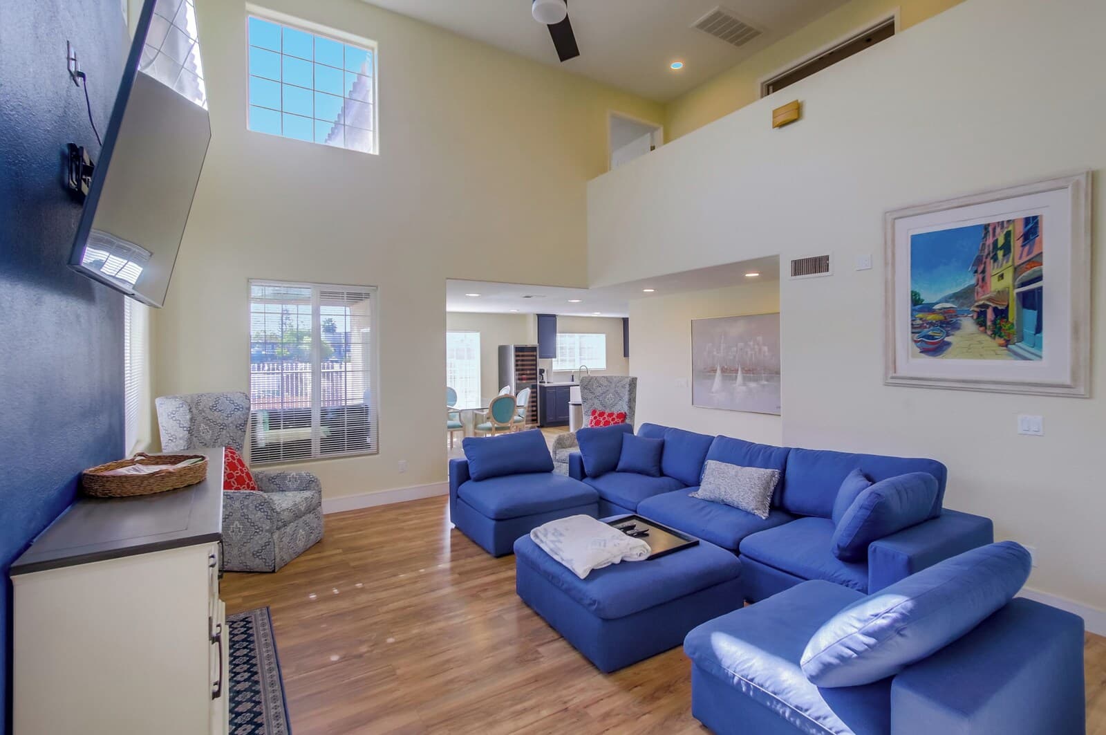 A spacious living room with a blue sectional sofa, a grey armchair, and a white cabinet. There is a painting on the wall and a window with blinds.