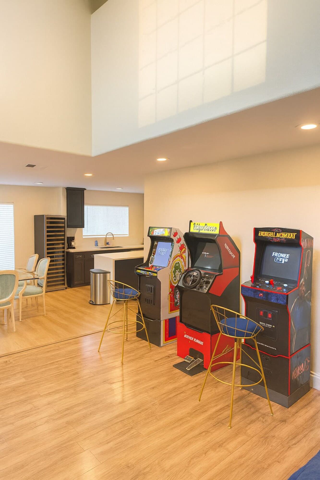 A room with wooden flooring, two arcade machines, a small kitchenette, and a seating area with chairs.
