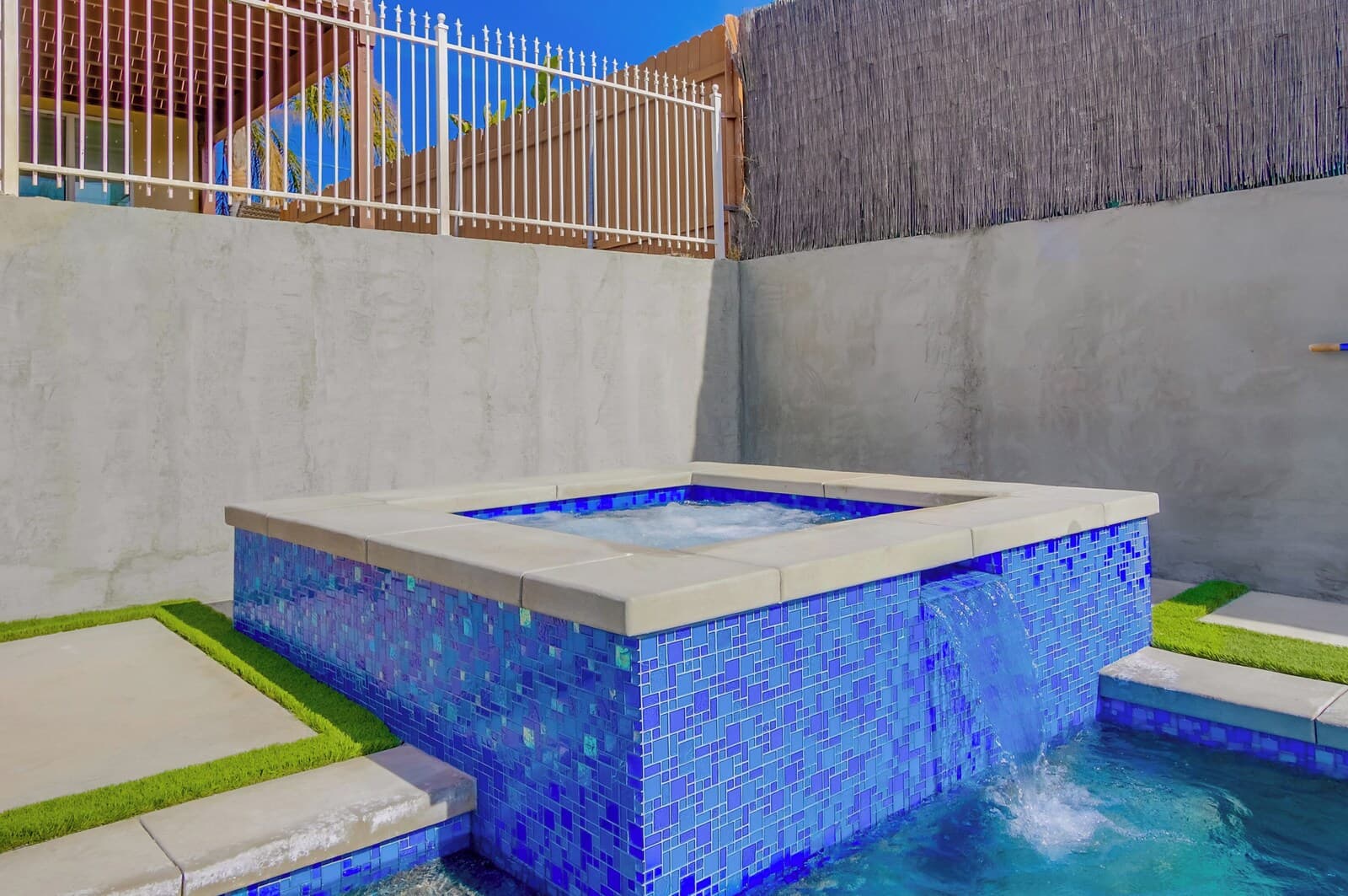 A square hot tub with blue mosaic tiles and a small waterfall feature, surrounded by a concrete deck and a grassy area.