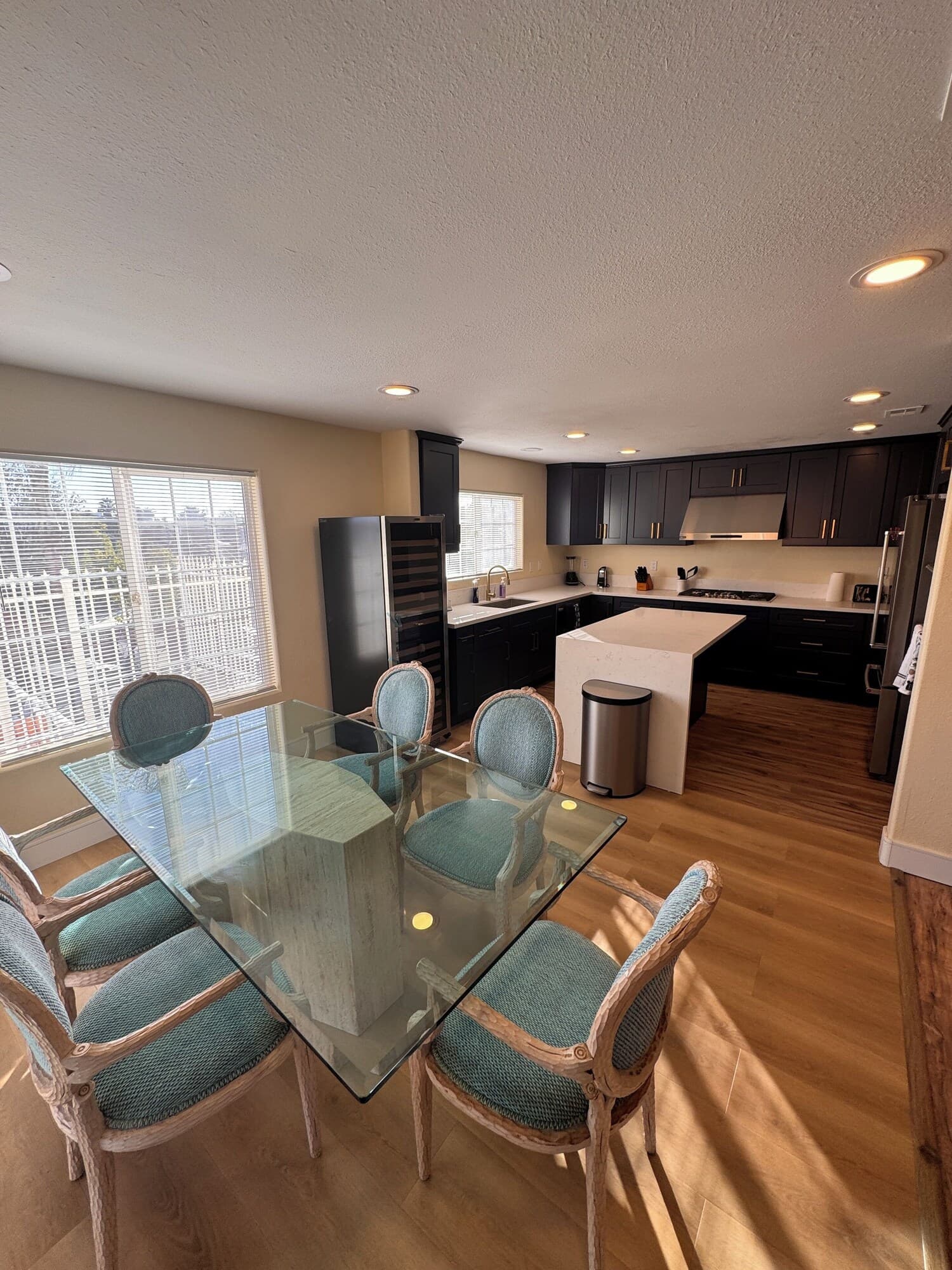 A modern kitchen with dark cabinets, a stainless steel range hood, and a glass dining table with four chairs.