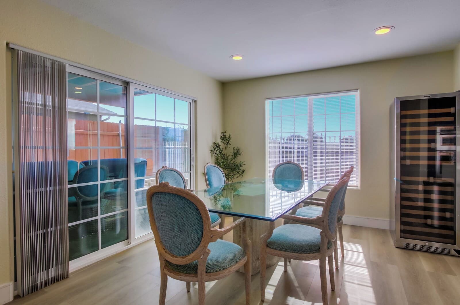 A dining area with a glass table and wooden chairs, large windows with blinds, and a sliding glass door.