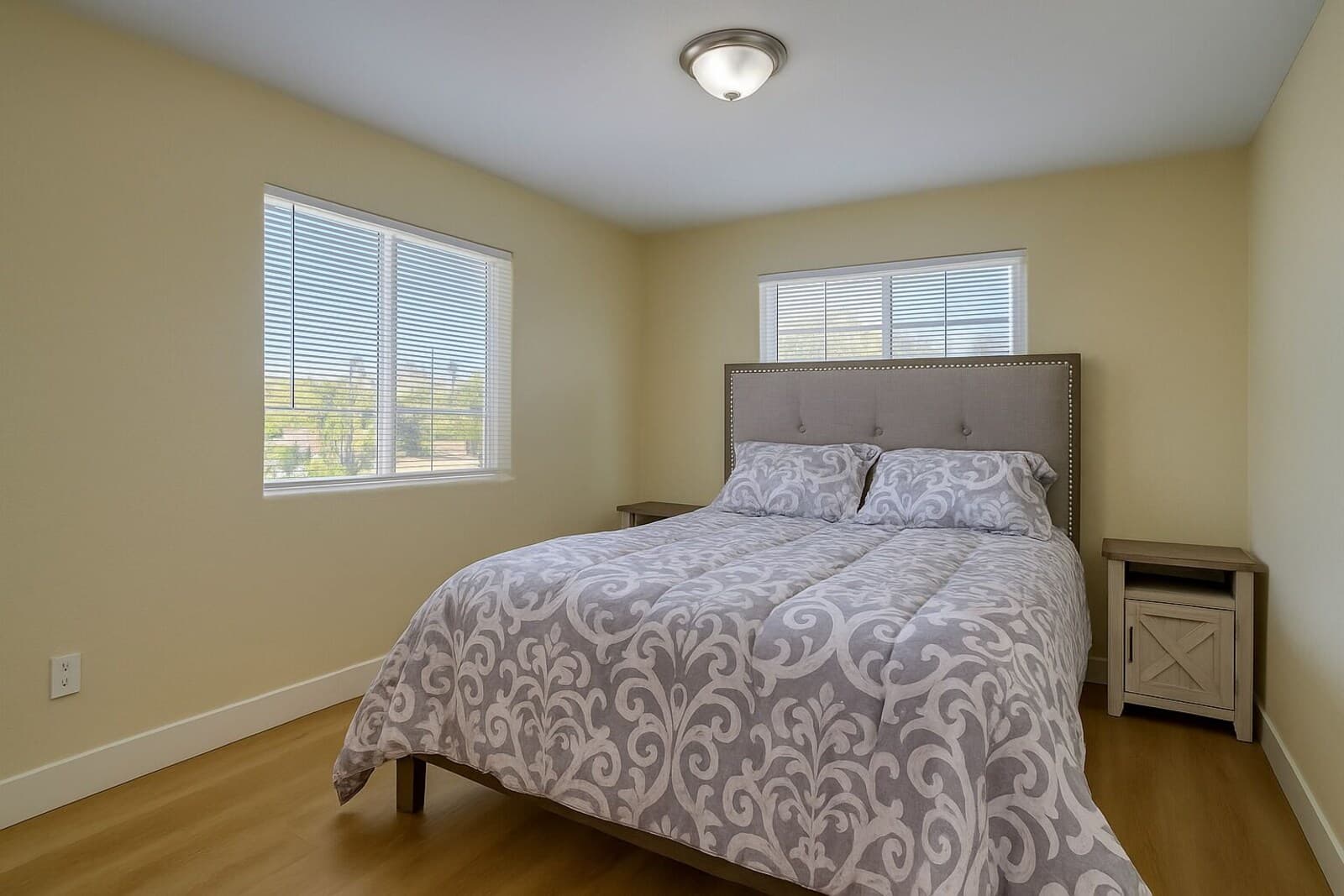 A bedroom with a bed, two nightstands, and two windows with blinds.