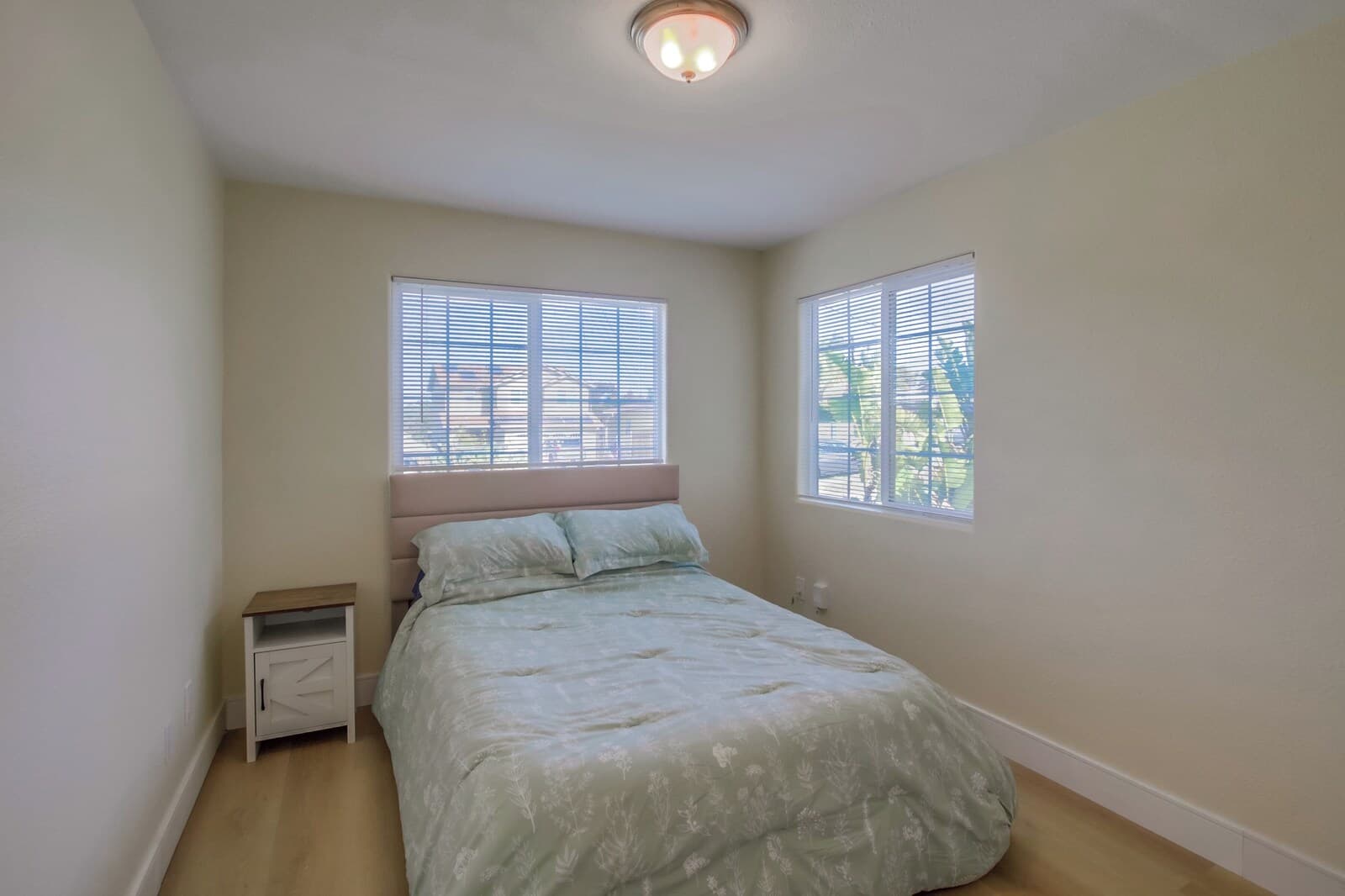 A bedroom with a bed, two windows with blinds, a nightstand, and a ceiling light.