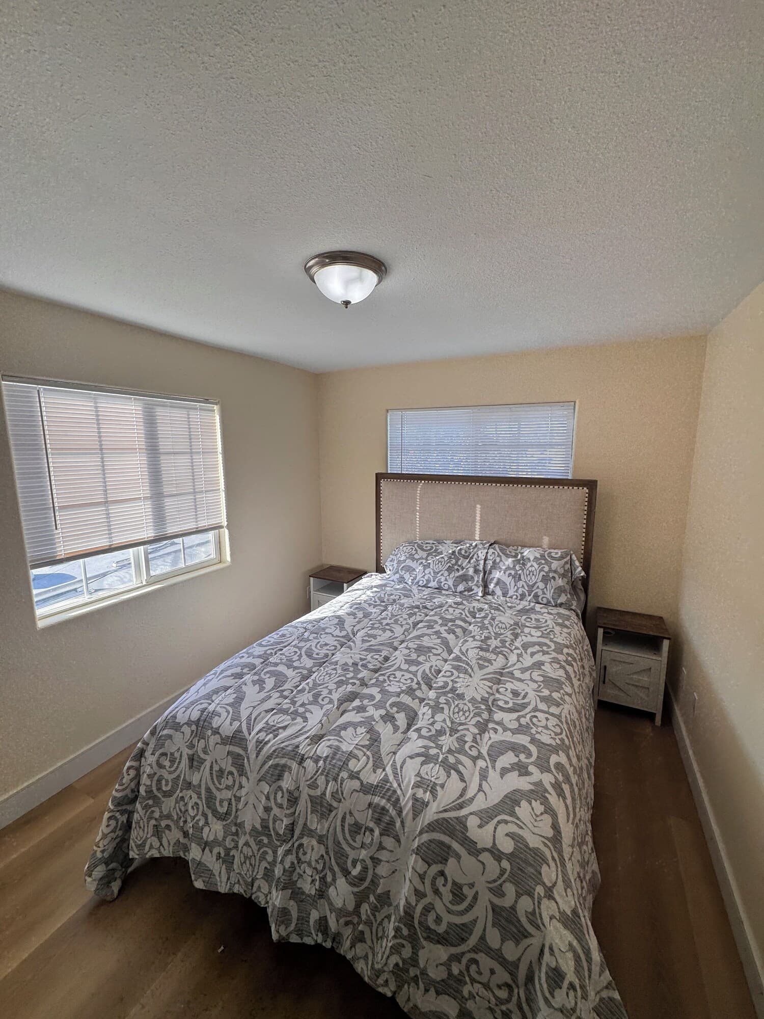 A bedroom with a bed, two nightstands, a ceiling light, and two windows with blinds.