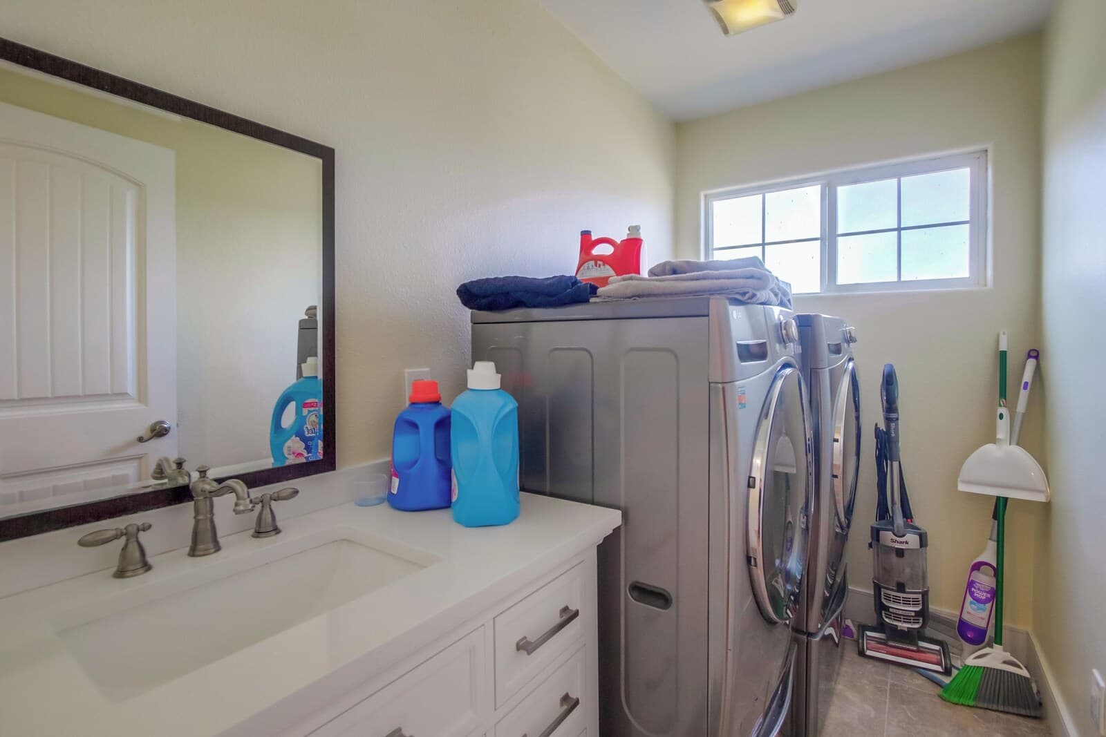 A bathroom with a white sink, two faucets, a mirror, a window, and a vacuum cleaner.