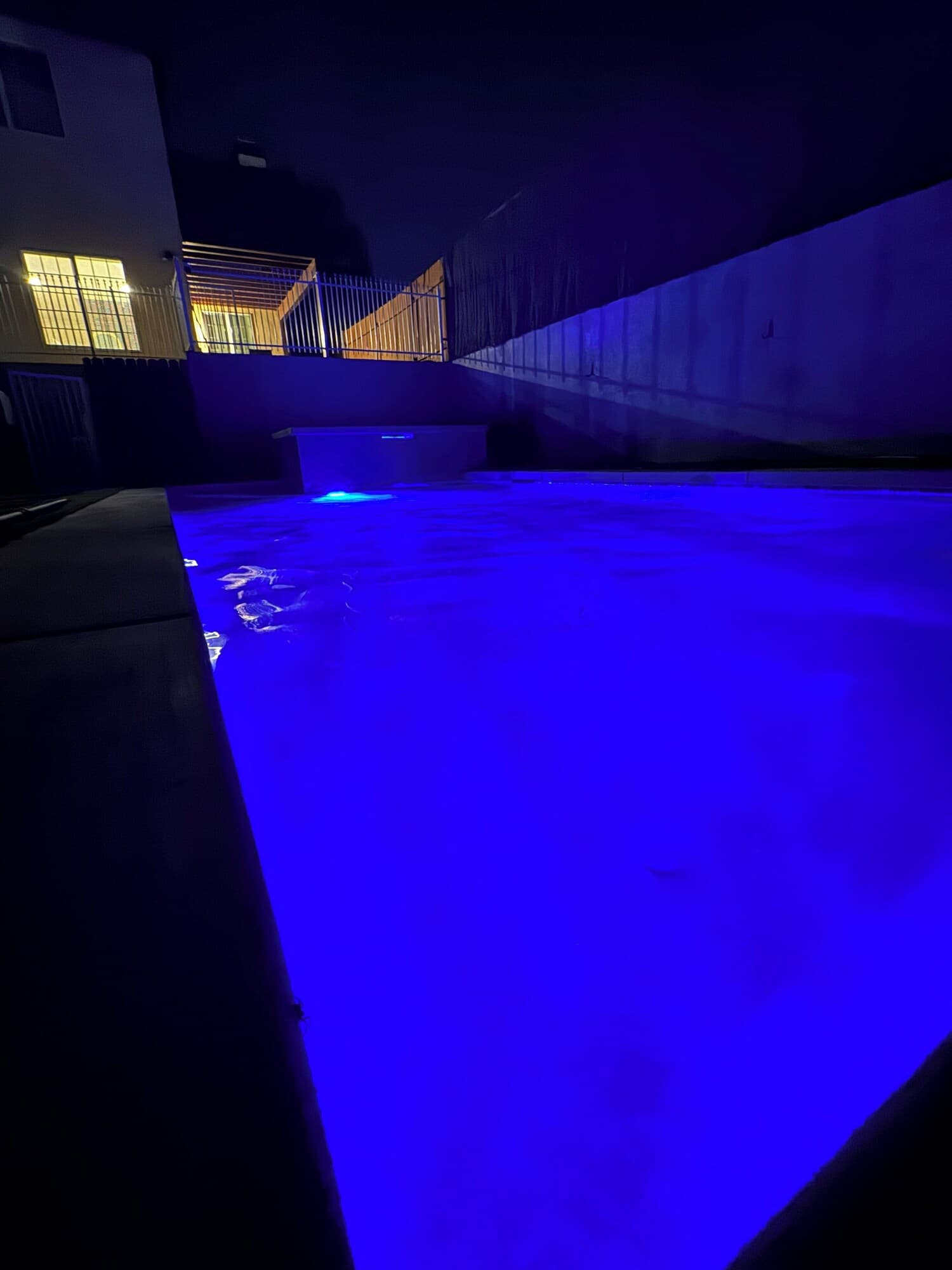 A swimming pool with a blue-lit water feature, surrounded by a concrete deck and a building with large windows.
