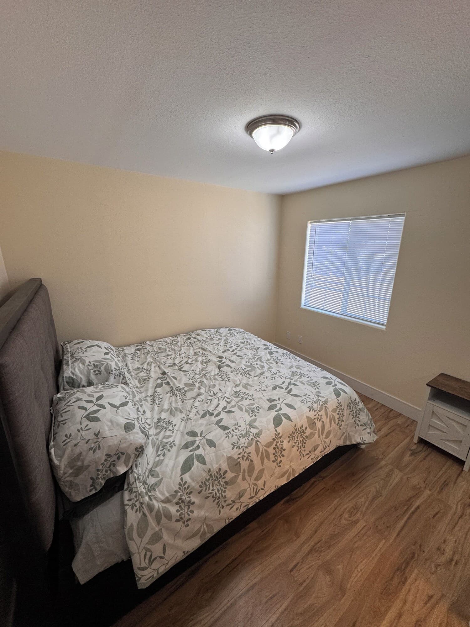 A bedroom with a bed, a window with blinds, a ceiling light, and a small wooden nightstand.